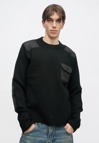 Melns džemperis ar teksturētām plecu un kabatu akcentiem, ribota apkakle un brīvs piegriezums. Valkāts ar gaiši zilām džinsām.