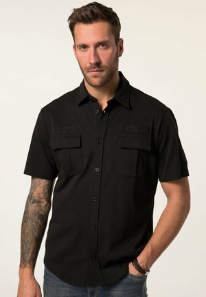 Man met kort bruin haar en baard, gekleed in een zwart overhemd met korte mouwen, twee borstzakken en tatoeages op zijn linkerarm.