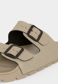 Sandale beige avec deux larges brides, chacune ornée d'une grande boucle foncée, et une semelle texturée sur un fond clair.