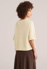 Femme aux cheveux bouclés portant un pull crème à manches courtes et une jupe marron, debout de profil devant un fond uni.