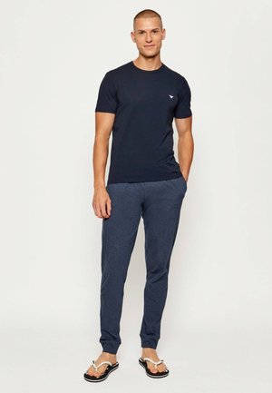 Uomo in piedi che indossa una maglietta nera aderente, pantaloni jogger blu scuro e infradito bianche e nere su sfondo bianco uniforme.