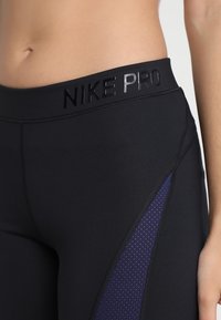 Černé legíny Nike Pro mají široký pas s vyraženým brandingem a fialové síťované detaily po stranách, které zlepšují prodyšnost a design.