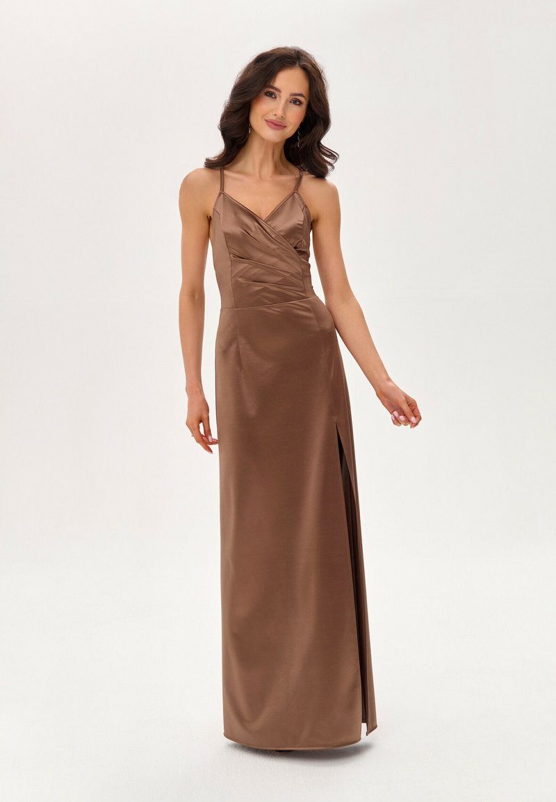 Žena s tmavými vlnitými vlasy, oblečená v hnědých saténových maxi šatech bez rukávů s bočním rozparkem, stojící na bílém pozadí.