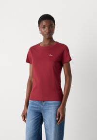 Maroon short-sleeve t-shirt made of soft cotton, featuring a round neck and a small embroidered logo on the chest, paired with blue jeans.