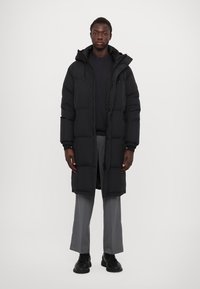Abrigo de plumas negro con capucha, con un diseño acolchado y cremallera frontal, llevado sobre una parte superior oscura y pantalones grises. Textura suave.
