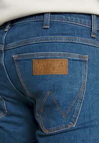 Jeans de denim azul com um retângulo de couro castanho apresentando o logótipo "Wrangler". Costuras expostas e um design distinto dos bolsos traseiros.
