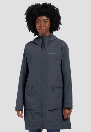 Kobieta ubrana w ciemnoszary, przeciwdeszczowy płaszcz z kapturem, z zamkiem błyskawicznym z przodu i dwoma kieszeniami na zamek, stojąca na tle jednolitego jasnoszarego tła.