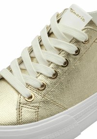 Metallic gold sneaker with white laces and eyelets. Features a textured surface and a white rubber sole. Brand name "Tamaris" visible on the tongue.