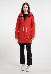 Giacca impermeabile rossa con un ampio cappuccio, coulisse regolabili, zip frontale e tasche laterali. Abbinata a pantaloni neri e sneaker bianche.