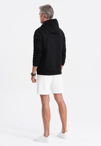 Sudadera negra con capucha alzada, puños y cinturilla acanalados, combinada con shorts blancos y zapatillas blancas, vista desde la espalda.