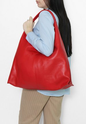 Donna con lunghi capelli scuri che indossa una camicia blu e pantaloni beige a righe sottili, che tiene una grande borsa a tracolla in pelle rossa.