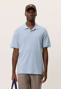 Ανοιχτό μπλε πόλο με κοντό γιακά, κοντά μανίκια και διακριτική λεπτομέρεια με λογότυπο. Φοράται με μπεζ παντελόνι και καφέ καπέλο.