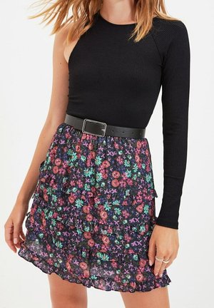 Frau trägt ein schwarzes, einseitig schulterfreies Langarmoberteil mit schwarzem Gürtel und einen mehrlagigen Minirock mit Blumenmuster.