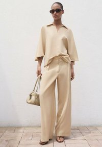 Beige weite Hosen und ein passendes, locker sitzendes Hemd mit Kragen. Eine weiche Handtasche und dunkle Sonnenbrillen runden den Look ab. Sanfte Stofftextur.
