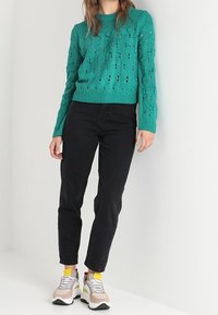 Tealstickad tröja med texturerat mönster, svarta jeans och flerfärgade sneakers med ljusbeige bas och vit sula.