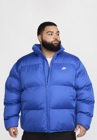 Mėlyna pūkinė striukė iš nailono, su aukštu apykakle, užtrauktuko uždarymu ir Nike logotipu kairiajame krūtinės šone; siuvinėta tekstūra ir per didelis fasonas.