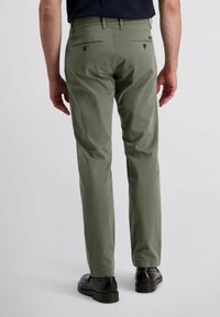 Olivgrüne Chinos mit geradem Schnitt, zwei hinteren Taschen mit Reißverschlüssen und einem glatten, leichten Stoff. Getragen mit schwarzen Schuhen.