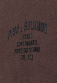 Hnědá bavlněná mikina s vyšitým černým textem: "PRM : STUDIOS (1990) COPENHAGEN PROCESS STUDIO 20/_23." Měkká textura, kulatý výstřih.