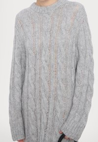 Pull gris clair en maille torsadée avec une coupe ample et un motif légèrement transparent, porté par une personne tenant un petit objet noir.