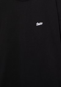 Sort stof med lille hvidt broderet "SND?"-logo på øverste venstre side.