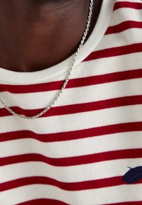 Maglietta a righe rosse e bianche con scollo rotondo, realizzata in tessuto morbido, con un piccolo dettaglio di design blu navy e una collana in argento.