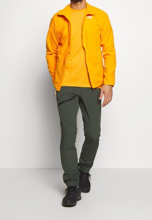 Hombre con chaqueta naranja brillante y camiseta, pantalones grises oscuros con cremallera y zapatos de senderismo negros sobre un fondo liso.