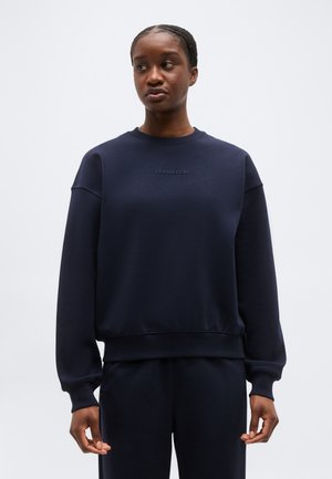 Γυναίκα με μπλε σκούρο φούτερ J.Lindeberg και αντίστοιχο παντελόνι, στέκεται κοιτάζοντας ελαφρώς προς τα αριστερά της, σε απλό φόντο.