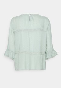 Mintgrünes Blusenshirt mit strukturiertem gepunktetem Muster, ausgestellt gefütterten Ärmeln und Spitzenakzenten, mit einem Knopfverschluss am Nacken hinten.