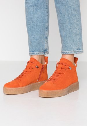 Pomarańczowe zamszowe trampki typu high-top z grubymi beżowymi podeszwami i zamkami błyskawicznymi po bokach, noszone z jasnoniebieskimi dżinsami.