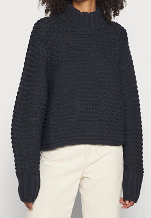 Personne portant un pull en maille côtelée noir à manches longues et col montant associé à un pantalon beige clair.