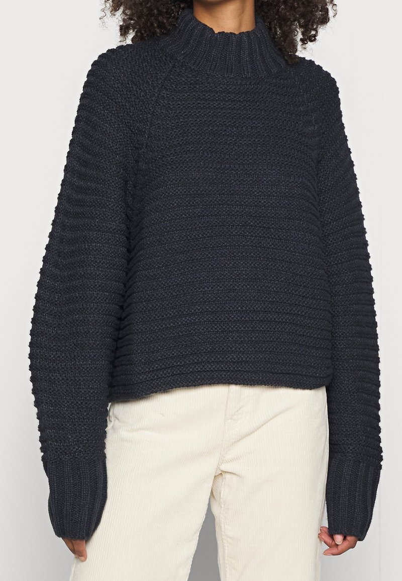 Personne portant un pull en maille côtelée noir à manches longues et col montant associé à un pantalon beige clair.