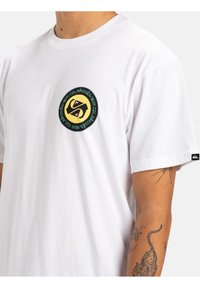 Camiseta blanca de algodón con un logo gráfico redondo en negro y amarillo, con texto verde en el pecho. Mangas cortas y diseño minimalista.