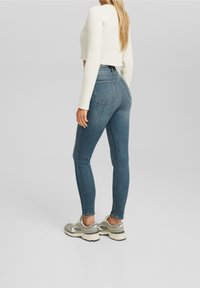 Vaqueros ajustados azules de mezclilla, con cintura alta, bolsillos traseros y una camiseta blanca corta texturizada acanalada. Combinado con zapatillas deportivas grises.