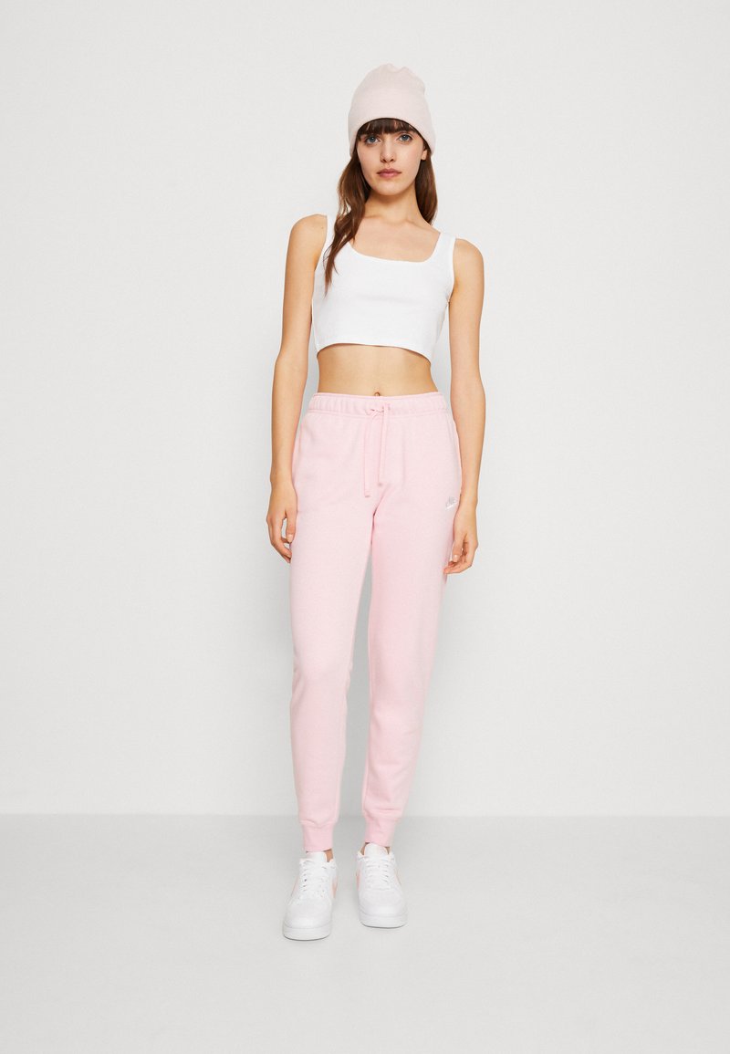 Jeune femme portant un crop top blanc, un jogging rose, un bonnet rose pâle et des baskets blanches, se tenant devant un fond blanc uni.