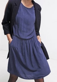 Mörkblå klänning med vita prickar, rund halsringning, elastisk midja och sidofickor; matchad med en mörk blazer.