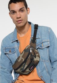 Kamouflage crossbody-väska i brun och grön med dragkedjeslutning, med svart hårdvara och justerbar rem, bärs över en jeansjacka.