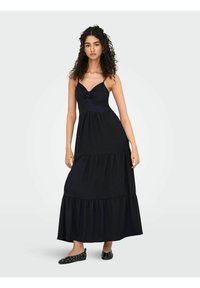 Robe maxi noire avec des bretelles spaghetti, un détail en nœud au niveau de la poitrine et un ourlet à volants en couches. Mannequin portant des ballerines noires avec des pois blancs.