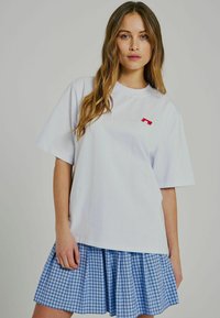 Weißes Baumwoll-T-Shirt mit kurzen Ärmeln, das ein kleines rotes Grafikmotiv zeigt. Kombiniert mit einem blau-weißen karierten, gefältelten Rock.