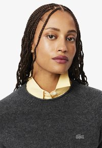 Maglione grigio a maglia con scollatura rotonda, indossato sopra una camicia gialla chiara con colletto. Caratterizzato da una sottile trama e un piccolo logo sul petto.