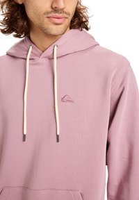 Sudadera rosa claro de tejido suave con cordón ajustable y un logo sutil en el pecho. Cuenta con un bolsillo delantero y puños acanalados.