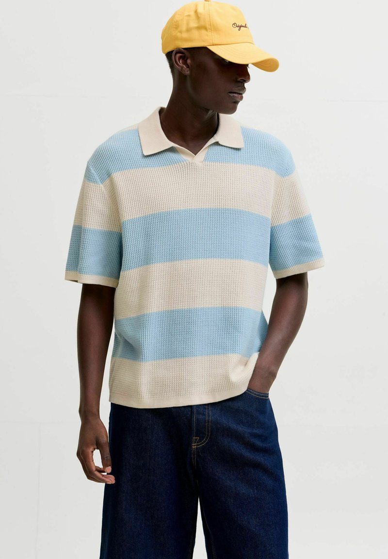 Homme portant une casquette jaune, un pull tricoté à manches courtes rayé bleu clair et beige, et un jean bleu foncé, debout avec une main dans la poche.