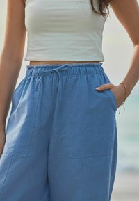 Pantaloni largi de culoare albastru deschis, confecționați dintr-un material moale, au o talie elastică cu șnur și buzunare laterale. Textură netedă și design simplu.