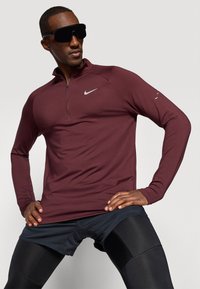 Bărbat îmbrăcat cu un tricou Nike cu mâneci lungi de culoare maro închis, pantaloni scurți sport negri și colanți, pozând cu mâinile în șolduri.