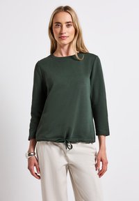 Felpa verde con scollo rotondo, maniche a tre quarti e dettaglio a fiocco all'orlo. Il tessuto sembra morbido e strutturato.