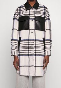 Checkered oversized coat with black leather accents, featuring large front pockets, buttons, and a contrasting collar. Soft fabric texture.