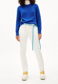 Blå stickad tröja, vita slim-fit jeans med ett flätat turkos bälte, och vita sneakers med gula snören. Slät textur, avslappnad design.