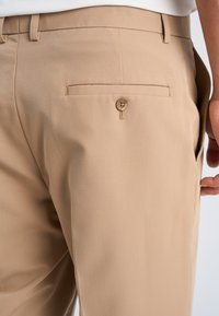 Beige skræddersyede bukser med bæltestropper og en knappet baglomme båret af en person, der står med en synlig højre hånd.