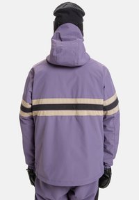 Giacca impermeabile viola con cappuccio, caratterizzata da strisce orizzontali nere e crema sulla schiena. Materiale in nylon testurizzato, vestibilità ampia.