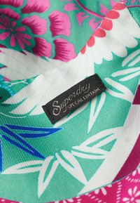 Tejido verde con patrones florales en rosa y blanco. Presenta una etiqueta negra con el texto "Superdry Edición Especial". Acabado texturizado.