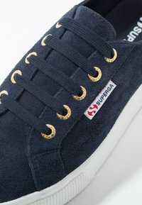 Navy blue suede sneaker featuring gold eyelets, dark laces, and a white rubber sole. Includes a red and white logo tag on the side.